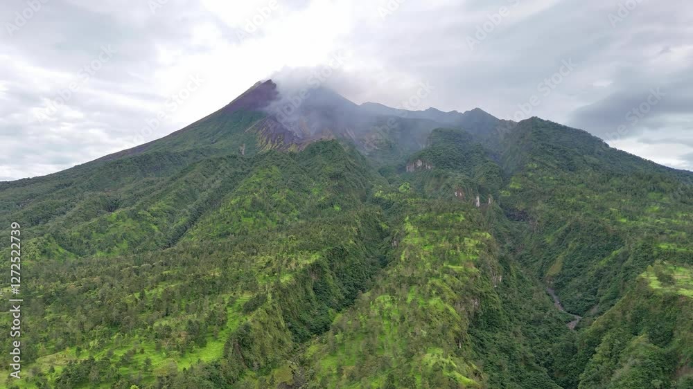 Drone view of a volcano emitting smoke from the crater at the top with green vegetation on the slopes. Merapi Volcano, Java, Indonesia. The most active volcano in Indonesia. 4K Aerial footage. Drone view of a volcano emitting smoke from the crater at the top with green vegetation on the slopes. Merapi Volcano, Java, Indonesia. The most active volcano in Indonesia. 4K Aerial footage.