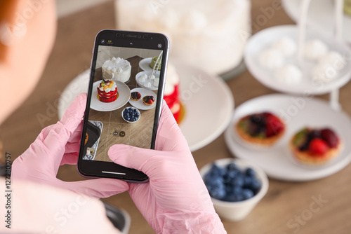 Fototapeta Naklejka Na Ścianę i Meble -  Female confectioner taking picture of cakes in kitchen, closeup