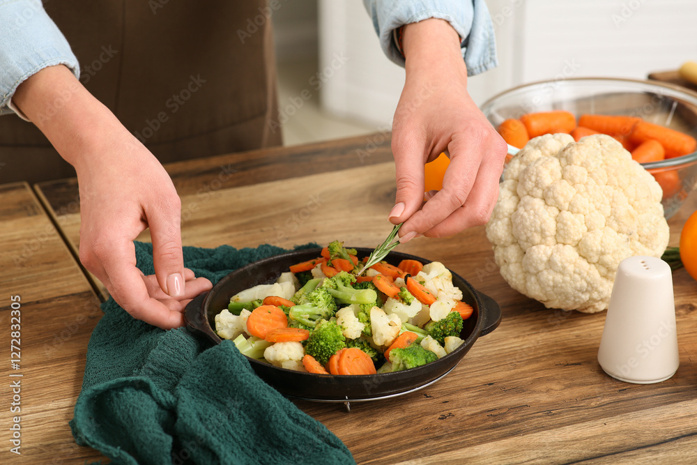 Young woman adding rosemary into frying pan with roasted vegetables in kitchen Young woman adding rosemary into frying pan with roasted vegetables in kitchen