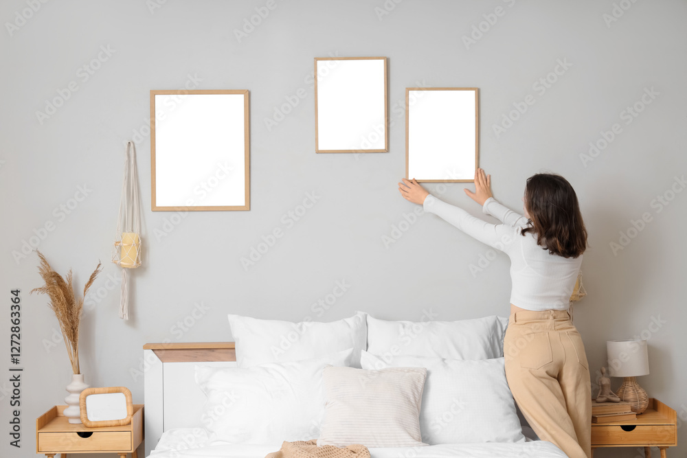 Young woman hanging blank frame on light wall in bedroom, back view