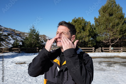 A man outdoors in a snowy landscape, wearing a black winter jacket, applying sunscreen to his face with closed eyes, protecting his skin from the sun at high altitude.
