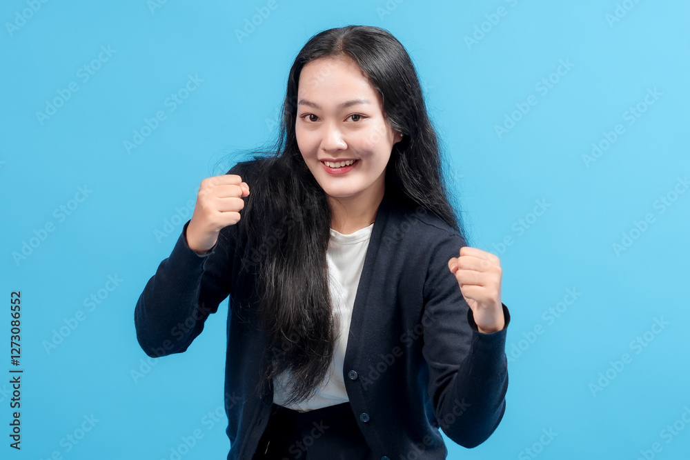Happy young Asian woman celebrating success with raised fists, wearing casual outfit, standing against blue background. Concept of achievement, victory, motivation, and positive energy. Happy young Asian woman celebrating success with raised fists, wearing casual outfit, standing against blue background. Concept of achievement, victory, motivation, and positive energy.