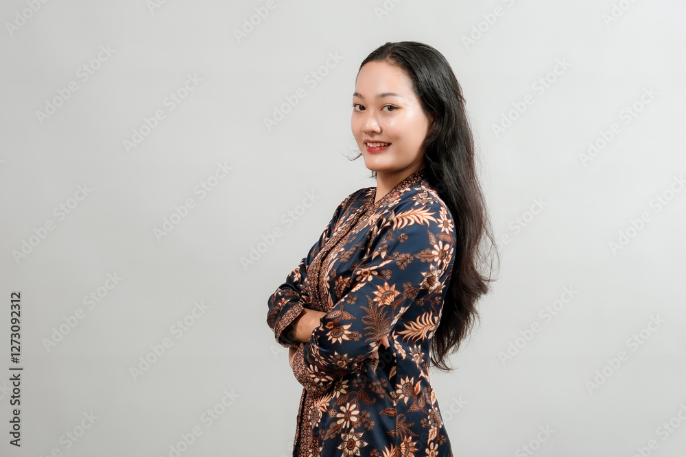 Confident young Asian woman wearing traditional batik dress, smiling with arms crossed, posing against neutral background. Confident young Asian woman wearing traditional batik dress, smiling with arms crossed, posing against neutral background.
