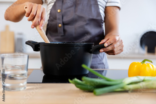 Woman chef using wooden spoon while stirring soup in a pot on induction stove, with fresh vegetables nearby in modern kitchen