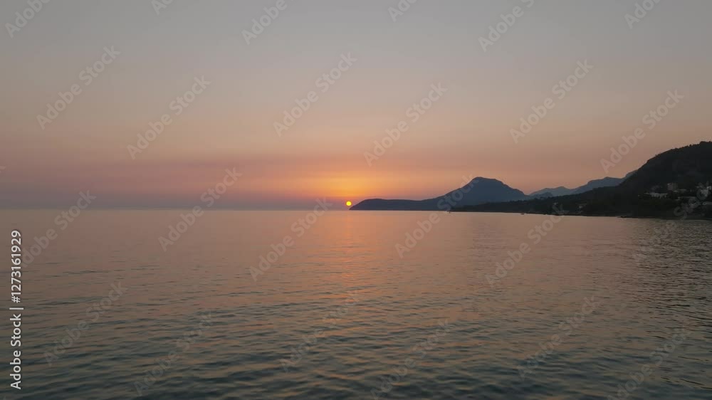 Drone view over the sea or ocean with a sunset at the center of the frame, with the silhouettes of mountains stretching into the sea Drone view over the sea or ocean with a sunset at the center of the frame, with the silhouettes of mountains stretching into the sea
