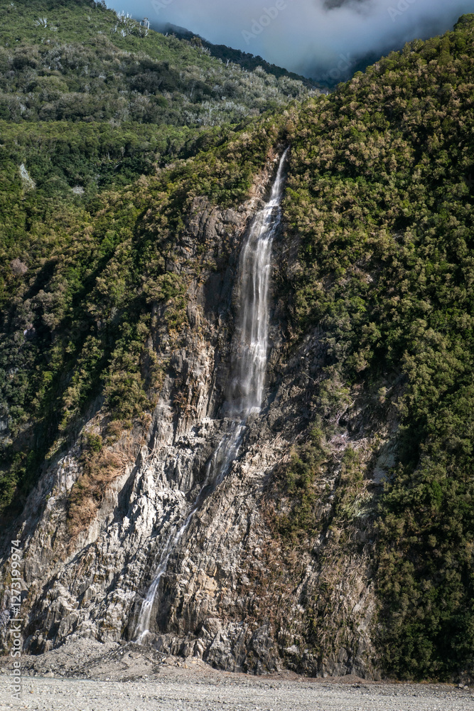Dramatic tall waterfall after heavy rain at Franz Josef Glacier surrounded with green nature with mist hovering in the background Dramatic tall waterfall after heavy rain at Franz Josef Glacier surrounded with green nature with mist hovering in the background