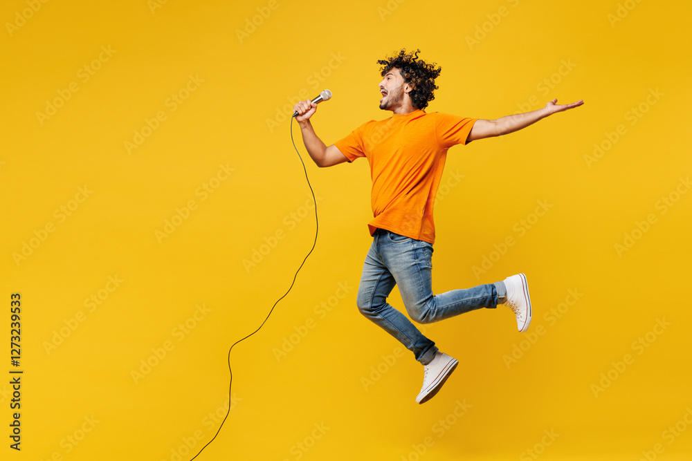 © ViDi Studio - Full body side view young Indian man he wearing orange t-shirt casual clothes jump high sing song in microphone at karaoke club isolated on plain yellow background studio portrait. Lifestyle concept.