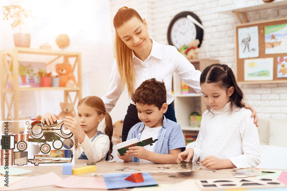 Fototapeta premium A young woman helps children fold parts from cardboard. This is their teacher. They have a lesson.