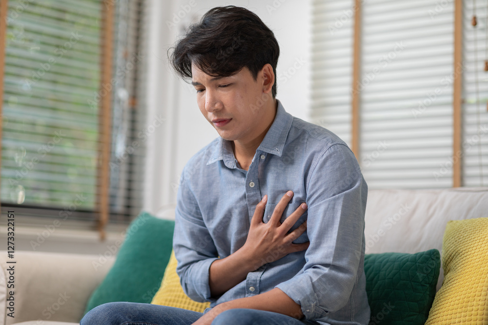man sitting on sofa, holding his chest with pained expression, possibly experiencing discomfort or heart related symptoms, in bright living room with cushions and natural light man sitting on sofa, holding his chest with pained expression, possibly experiencing discomfort or heart related symptoms, in bright living room with cushions and natural light