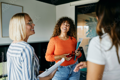 Friendly Office Interaction Among Diverse Women in a Modern Workspace. The three women are talking together, smiling, with notebooks in their arms