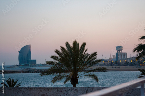 Palm tree silhouette against the twilight sky, with Barcelona landmarks visible in the distance. Coastal scene, cityscape, and a tranquil atmosphere.
