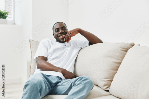 Smiling African man relaxing on a beige sofa, dressed in a casual white t shirt and blue jeans, exuding happiness in a bright, minimalist living room