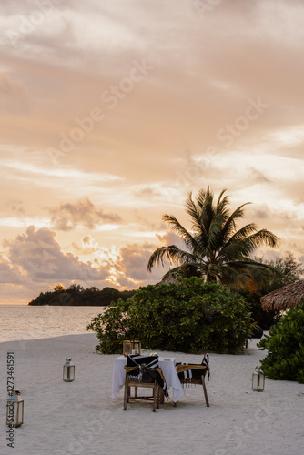 Fototapeta Naklejka Na Ścianę i Meble -  A beautifully set dining table on a sandy beach at sunset, overlooking the ocean and a distant island. A serene and romantic setting for an intimate dinner by the sea in Maldives