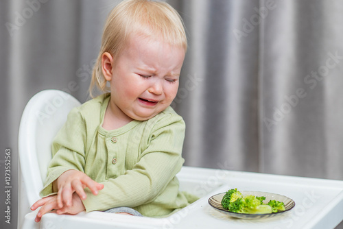 Cute baby refuses to eat broccoli at the kitchen table