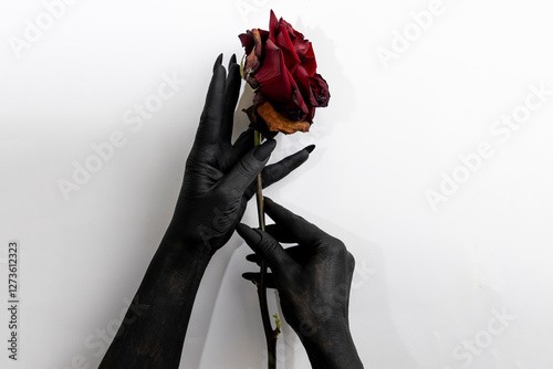 The black hands with long nails hold a rose and make it dry. Concept photo with woman hands painted in black paint with hold a red flower. 