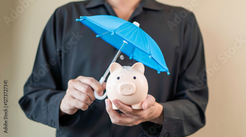 Obraz na płótnie financial security, savings protection, and money management, a person holding a piggy bank under a small blue umbrella. insurance, banking, and responsible financial planning. w salonie