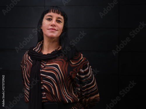 A confident woman in a stylish brown patterned blouse poses playfully against a sleek dark background, showcasing modern fashion and natural charm in an urban setting