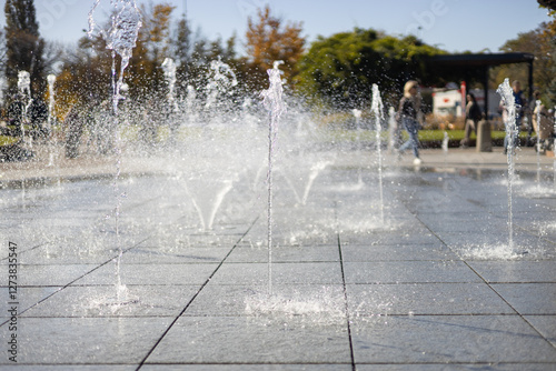 Obraz na płótnie Water droplets spread from the fountain in the air. Splashing water from a fountain in the park. Vertical fountain jets in the sidewalk on the square in the city park. w salonie