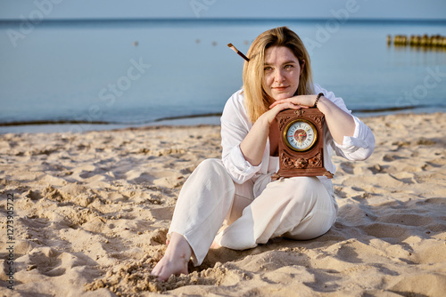 Young woman sits on sandy beach holding ornate clock, embodying themes of time and reflection. Serene seascape and soft light evoke sense of tranquility and introspection photo