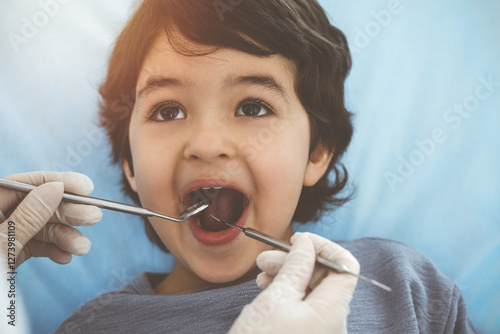 Cute arab boy sitting at dental chair with open mouth during oral checking up with doctor. Visiting sunny dentist office