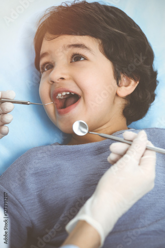 Cute arab boy sitting at dental chair with open mouth during oral checking up with doctor. Visiting sunny dentist office