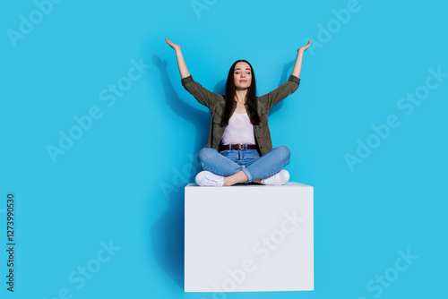 Young woman sitting on a white block with arms raised seated cross-legged against a bright blue background