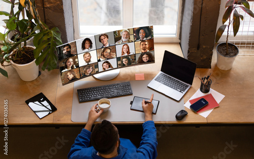 Wallpaper Mural Overhead view of remote worker in blue jacket holding coffee, engaging in online meeting with large virtual team displayed on monitor against cozy home office. Concept of teleworking, networking. Torontodigital.ca