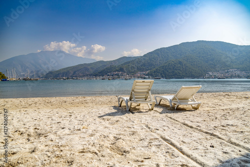 Fototapeta Naklejka Na Ścianę i Meble -   Sun loungers on the white sand of seashore with panorama of the city and mountains.  Karagozler. Fethiye, Mugla, Turkey.