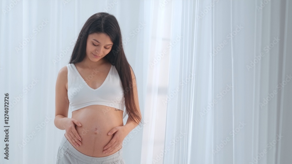 Young happy Asian pregnant woman posing in studio with white curtains in background Young happy Asian pregnant woman posing in studio with white curtains in background