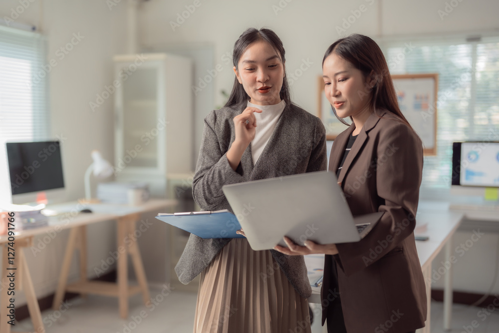 Two businesswomen engage in a collaborative discussion, using a laptop and clipboard in a bright, modern office setting. They are focused on teamwork and productivity in a professional environment Two businesswomen engage in a collaborative discussion, using a laptop and clipboard in a bright, modern office setting. They are focused on teamwork and productivity in a professional environment