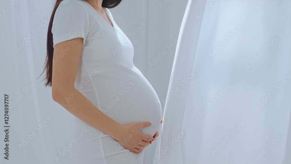 Young happy Asian pregnant woman posing in studio with white curtains in background Young happy Asian pregnant woman posing in studio with white curtains in background