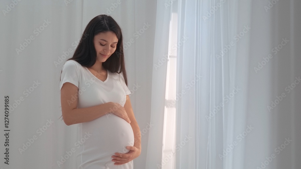 Young happy Asian pregnant woman posing in studio with white curtains in background Young happy Asian pregnant woman posing in studio with white curtains in background