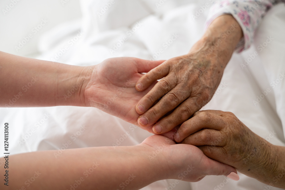 A young womans hands gently hold the hands of an elderly female patient in a hospital bed, symbolizing care, support, and compassion. A young womans hands gently hold the hands of an elderly female patient in a hospital bed, symbolizing care, support, and compassion.