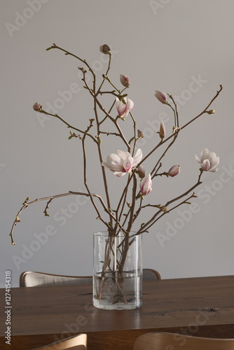 A magnolia branch in a clear glass vase rests on an oak stump, embodying spring in a Japandi interior. The soft blooms and warm wood create a serene, minimalist atmosphere. .