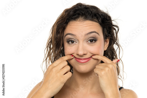 Young woman with makeup, pressing fingers against cheeks to create exaggerated smile. Playful expression exploring facial emotions. Studio portrait on white background photo