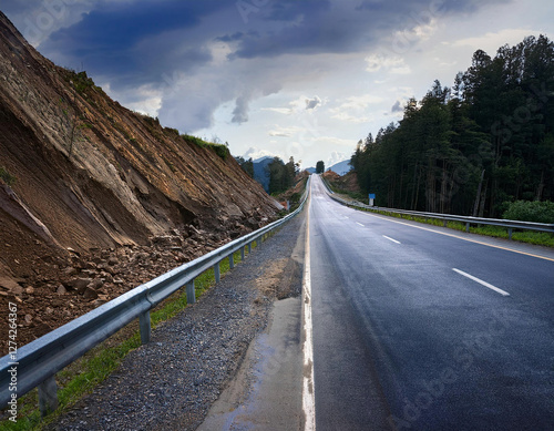 A long, wet asphalt road stretches into the distance, surrounded by a forest on one side and a rocky landslide area on the other, under a cloudy sky