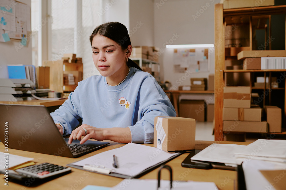 Woman working at desk, typing on laptop and reviewing documents in an office filled with cardboard boxes