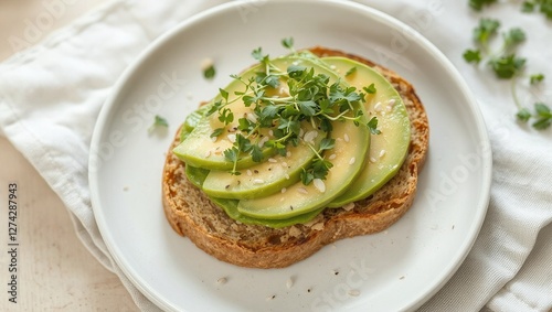 A beautifully styled avocado toast on whole-grain bread, topped with microgreens and sesame seeds. The plate rests on a neutral linen napkin with soft, natural lighting emphasizing the textures and co