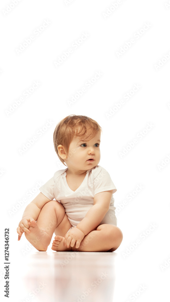 Baby, dressed white onesie, sitting cross-legged while looking sideways with focused expression against white background. Concept of childhood, born and growth, awareness, observation, early cognition