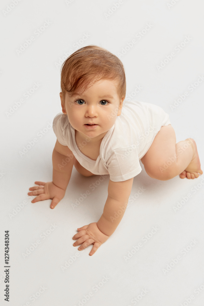 Aerial view of playful baby, dressed white onesie, crawling forward with bright eyes focused ahead against white studio background. Concept of movement, development, curiosity, determination. Aerial view of playful baby, dressed white onesie, crawling forward with bright eyes focused ahead against white studio background. Concept of movement, development, curiosity, determination.