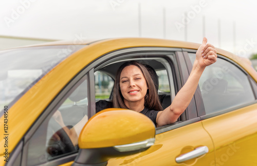 Obraz na płótnie A young woman with long brown hair and a bright smile sits in the driver's seat of a yellow car, giving a thumbs-up gesture through the open window with a confident and positive expression. w salonie