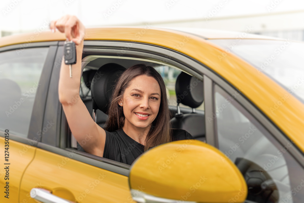 A young woman with long brown hair and a bright smile sits in the drivers seat of a yellow car, happily holding up a set of car keys through the open window.