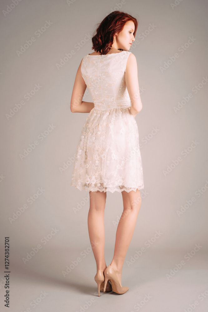 Fototapeta premium Studio portrait of attractive young ginger woman in double layer lace dress on high heels. Printed midi sleeveless white gown. Natural makeup and hairstyle. Modern bridal fashion, rear view