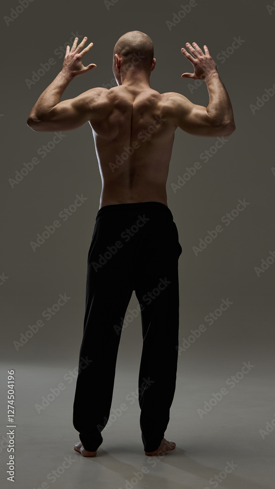 Full length, back view of ale model raising hands in expressive motion showcasing defined back muscles against deep brown background. Concept of beauty, fashion, style, masculinity. Full length, back view of ale model raising hands in expressive motion showcasing defined back muscles against deep brown background. Concept of beauty, fashion, style, masculinity.