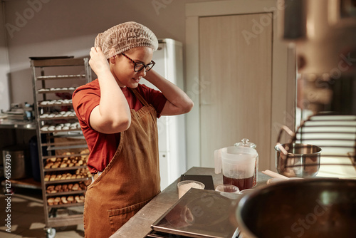 Medium shot of young female baker in apron adjusting hairnet preparing to mix ingredients for dough while standing at counter in bakehouse kitchen