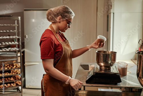 Obraz na plátně Side view of young female baker wearing protective hairnet scaling cocoa powder