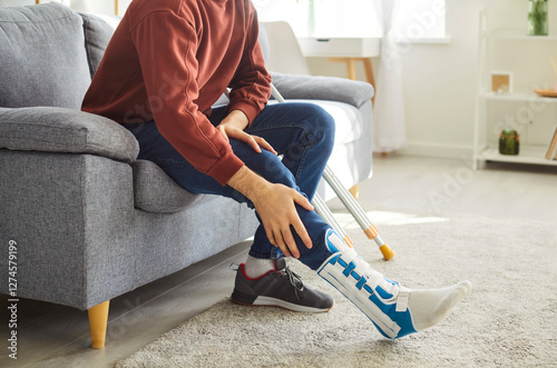 Close-up of handicapped man in casual clothes with leg in cast with crutch sitting on sofa at home in rehabilitation and recovering after having fractured limb. Recovery, accident and rehab concept.