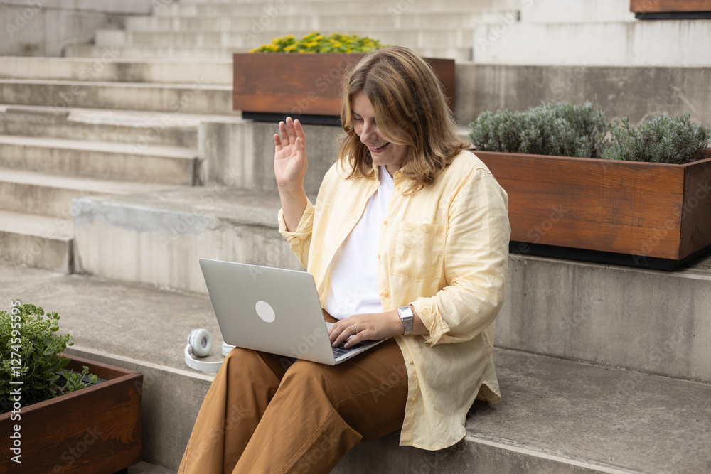 Happy 30s woman doing business on laptop and smiles. Young woman enjoy her work outdoor. Remote work, freelance. Young pretty female talking on video call and seating on stairs