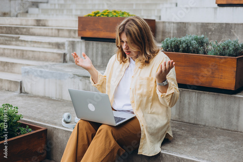 Angry shocked young woman looking at laptop screen, sitting on steps outdoors, she reading bad news in email or message, financial problem. Negative emotion.