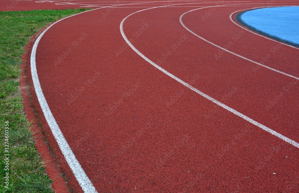 Fototapeta premium Red running track curve with blue sports field in right side and green grass in left side, sunny day